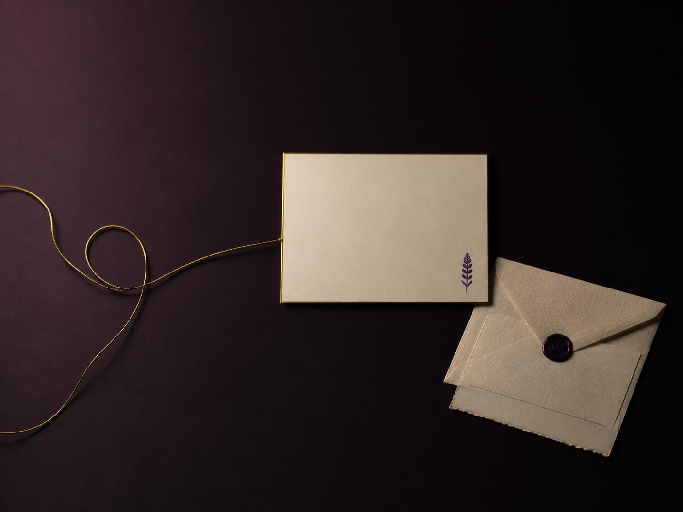 A single finished ivory card with an antique-gold edge and a small violet detail resting on deep aubergine, with a gold thread arriving at it beside a wax-sealed parchment envelope